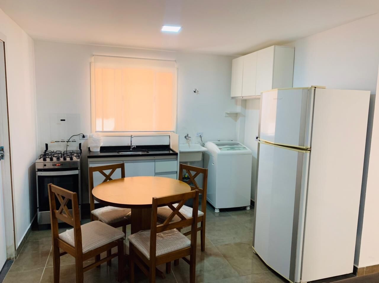 A compact kitchen with a stove, sink, and refrigerator, a round wooden table with chairs, and a window with blinds.