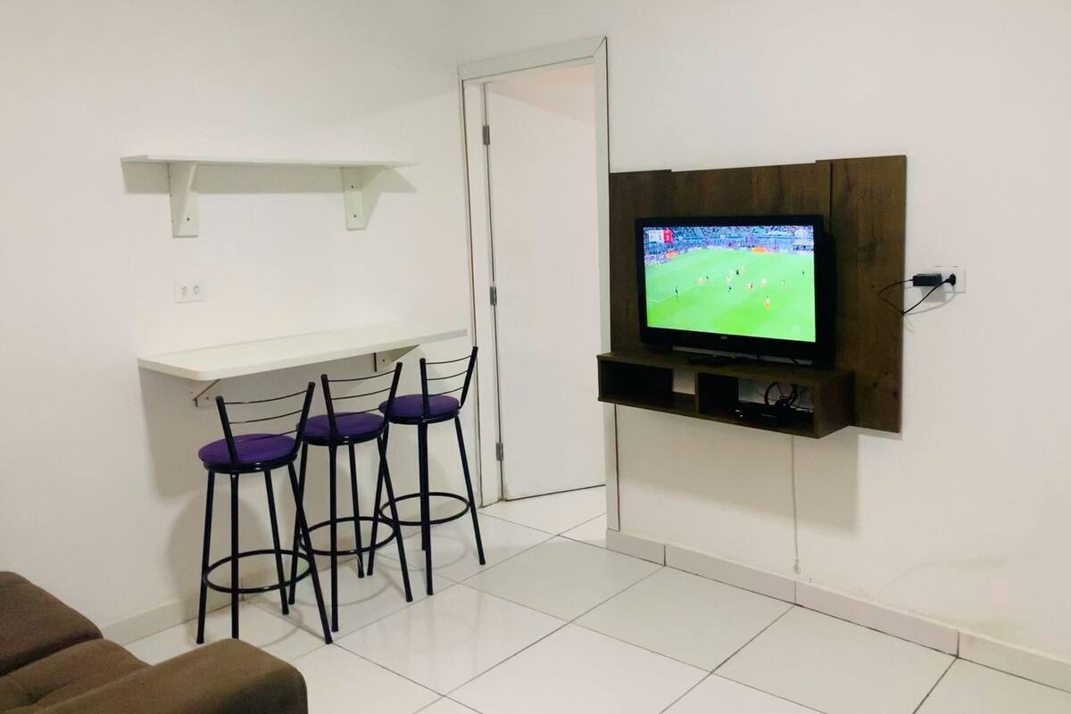 Un salón con un televisor montado en la pared, un estante blanco y tres sillones morados.