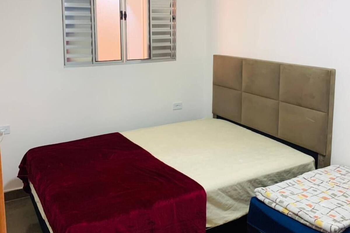 Un dormitorio con una cama individual, una cama más pequeña, una ventana con persianas y una pared blanca lisa.