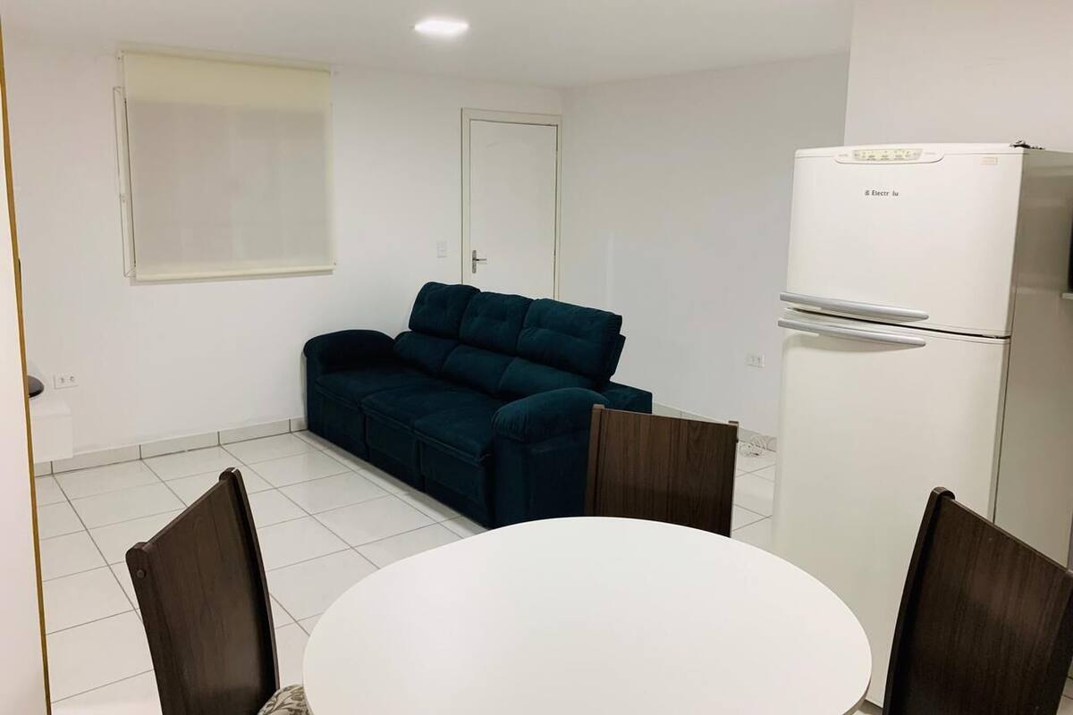 Una habitación pequeña y limpia con una mesa blanca y cuatro sillas, un sofá azul oscuro, un refrigerador blanco y una ventana con persianas.