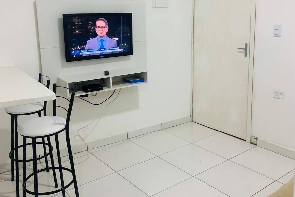 Una habitación con televisión montada en la pared, una mesa blanca con dos sillas y una puerta cerrada.