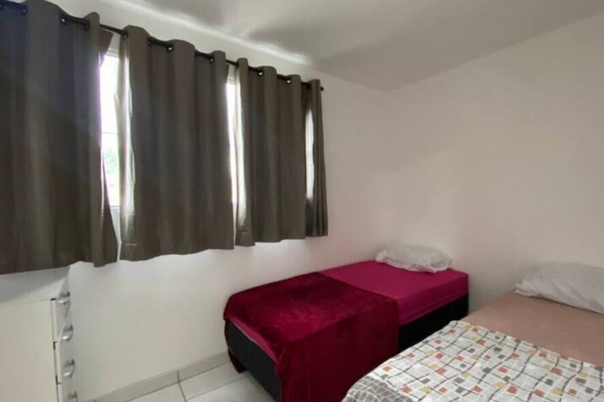 Una habitación pequeña con una cama individual, colcha roja, almohada blanca y ventana con cortinas.