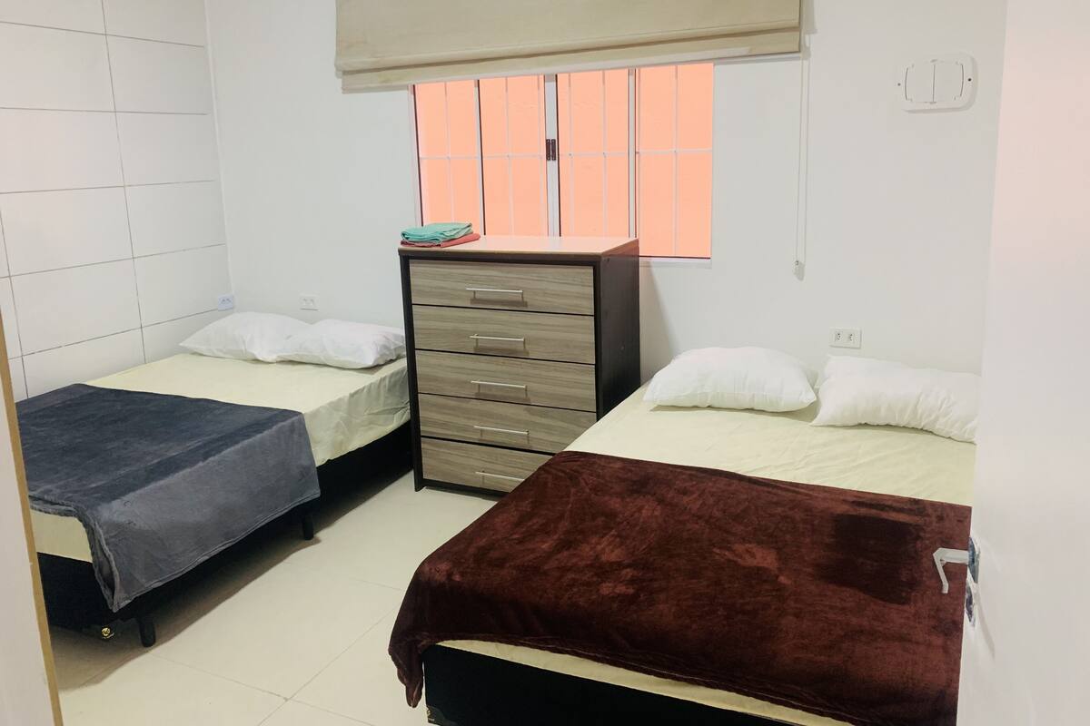 Habitación tipo dormitorio con dos camas individuales, un aparador de madera, una ventana con persianas y una pared con baldosas.