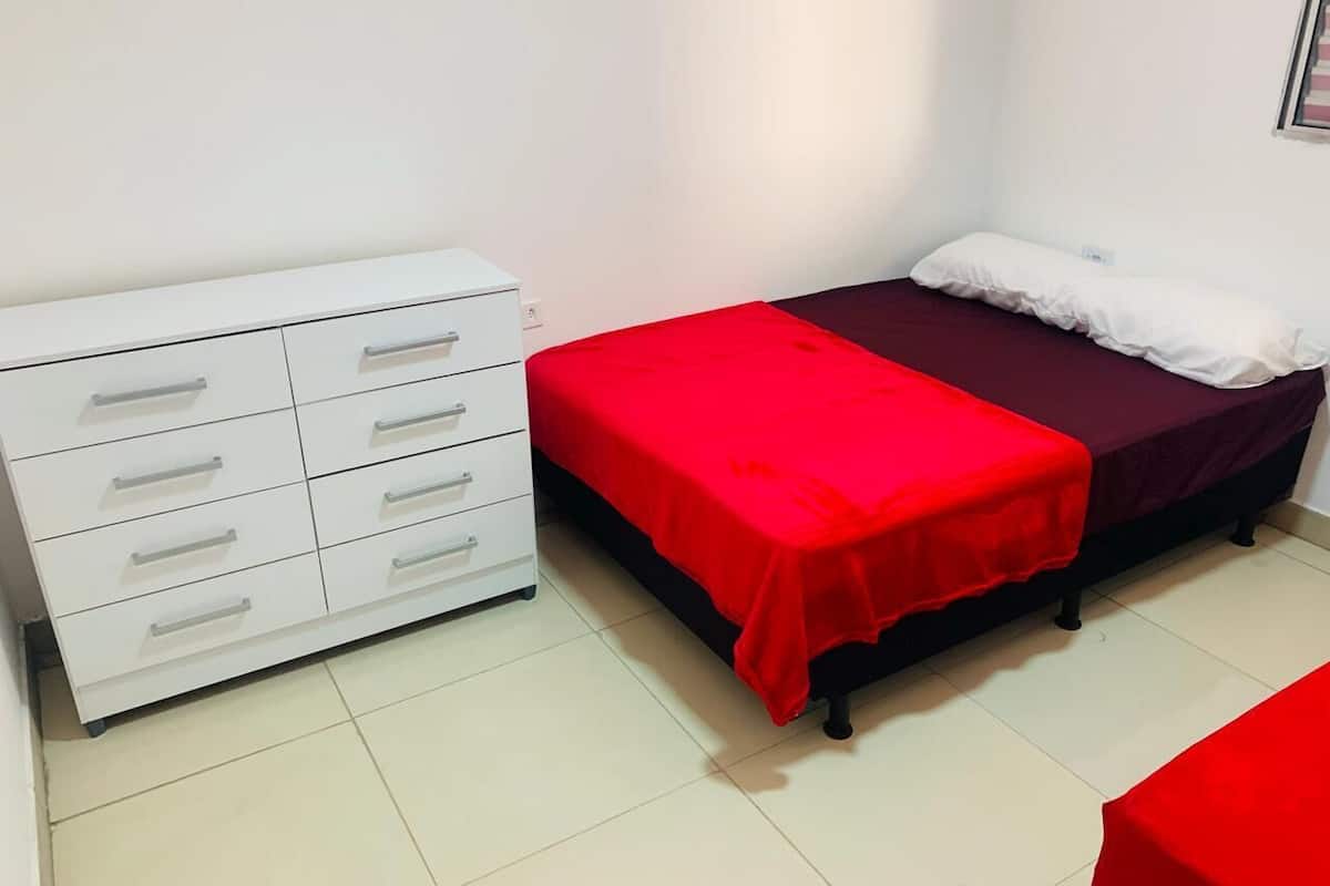 Una habitación pequeña y limpia con una cama, un aparador blanco y una colcha roja.