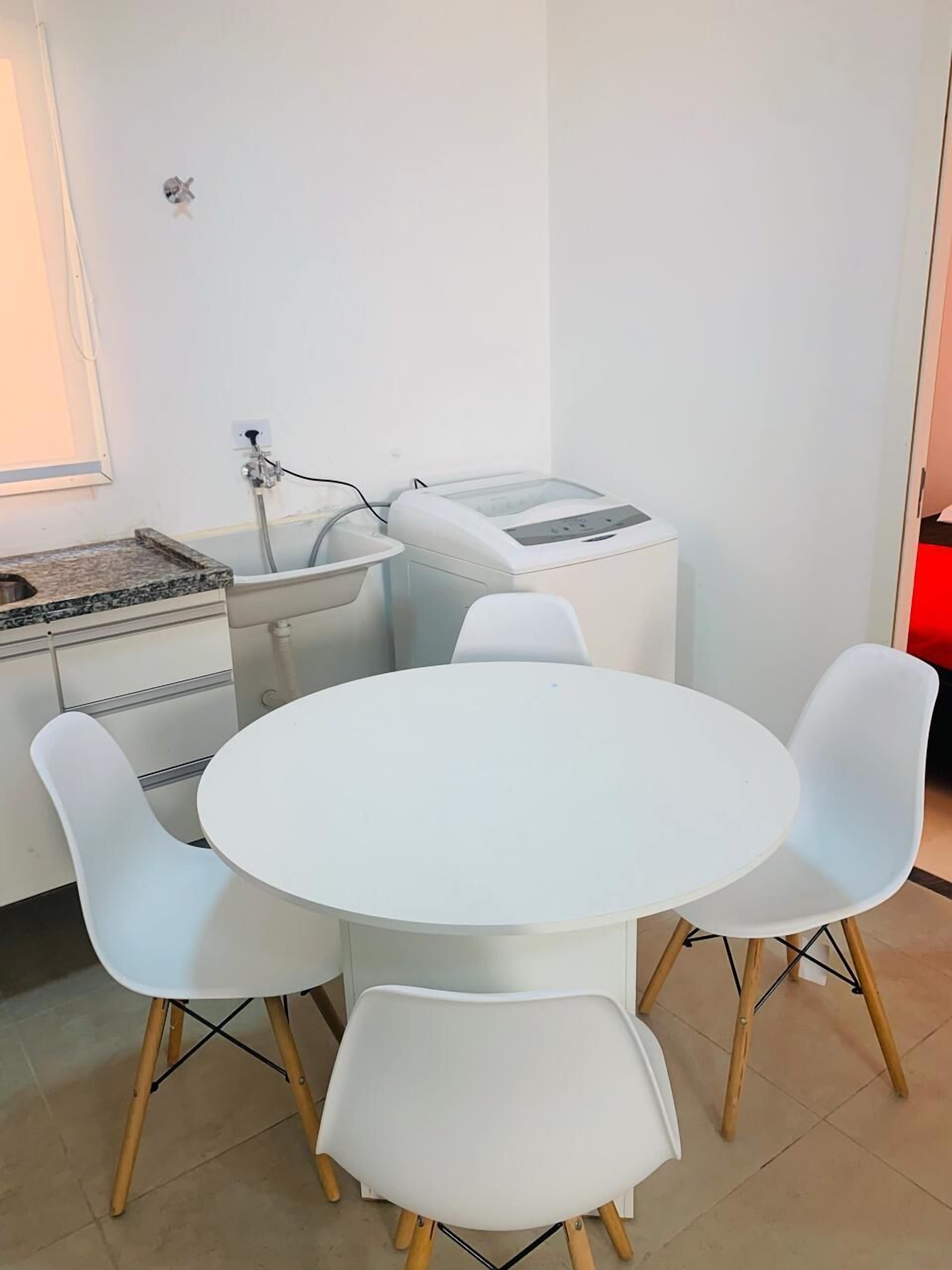 A small, modern room with a white round table and four white chairs with wooden legs. There is a washing machine and a small sink with a hose.