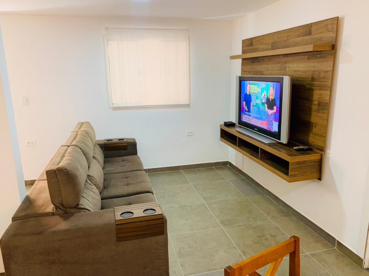 Un salon avec un canapé, un téléviseur fixé sur une étagère en bois, une fenêtre avec des stores et un sol carrelé.