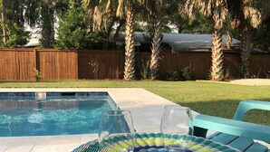 Une piscine avec une clôture en bois en arrière-plan, une cour gazonnée et une chaise bleue avec un set de table en fibres tressées.