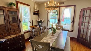 A dining area with a wooden table, chairs, a chandelier, and a view of the outdoors through large windows.