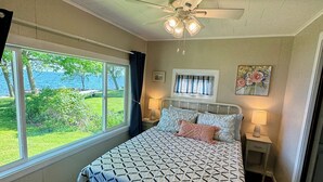 A bedroom with a bed, a ceiling fan, a window with a view of the outdoors, a nightstand, and a wall-mounted lamp.