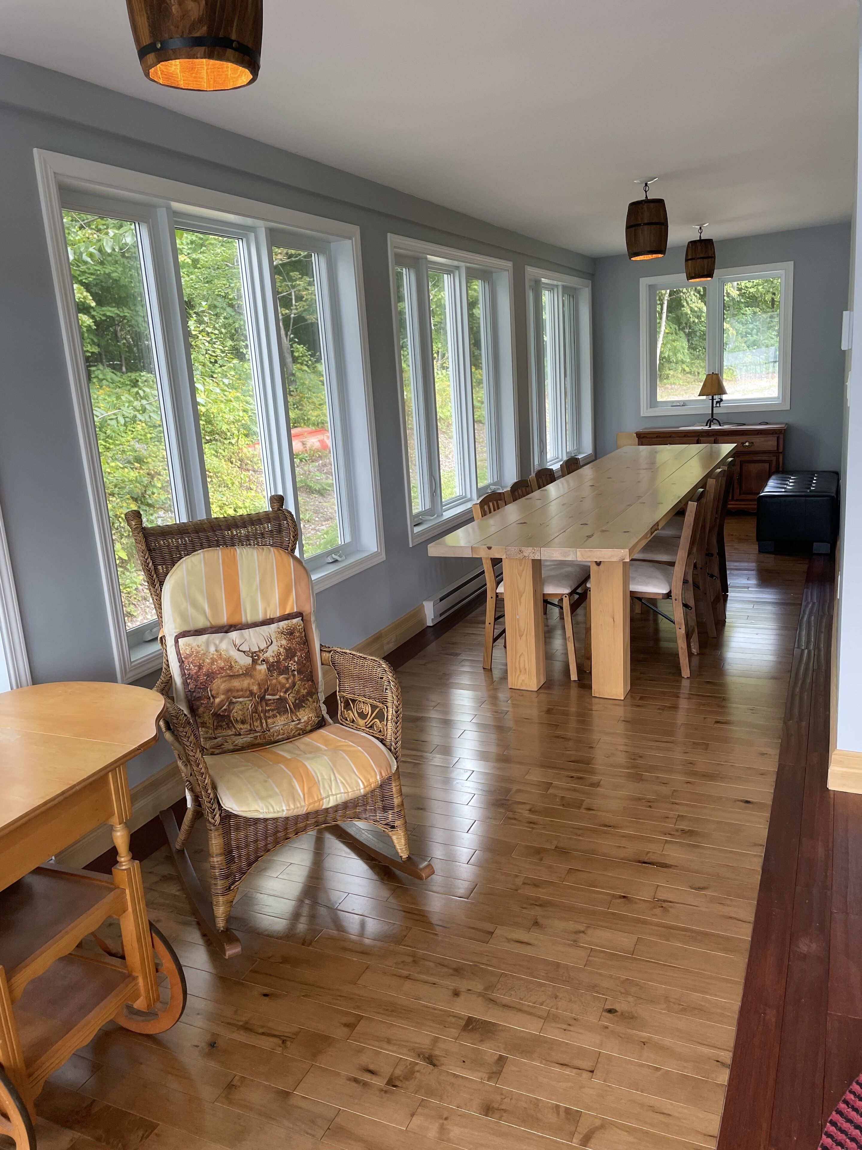 A dining area with a long wooden table, wicker chairs, and large windows offering a view of greenery.
