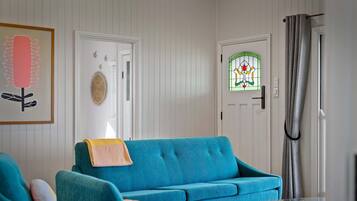 Een woonkamer met een blauwgroene bank, een glazen salontafel en een deur met een raam in glas-in-lood met groene details.