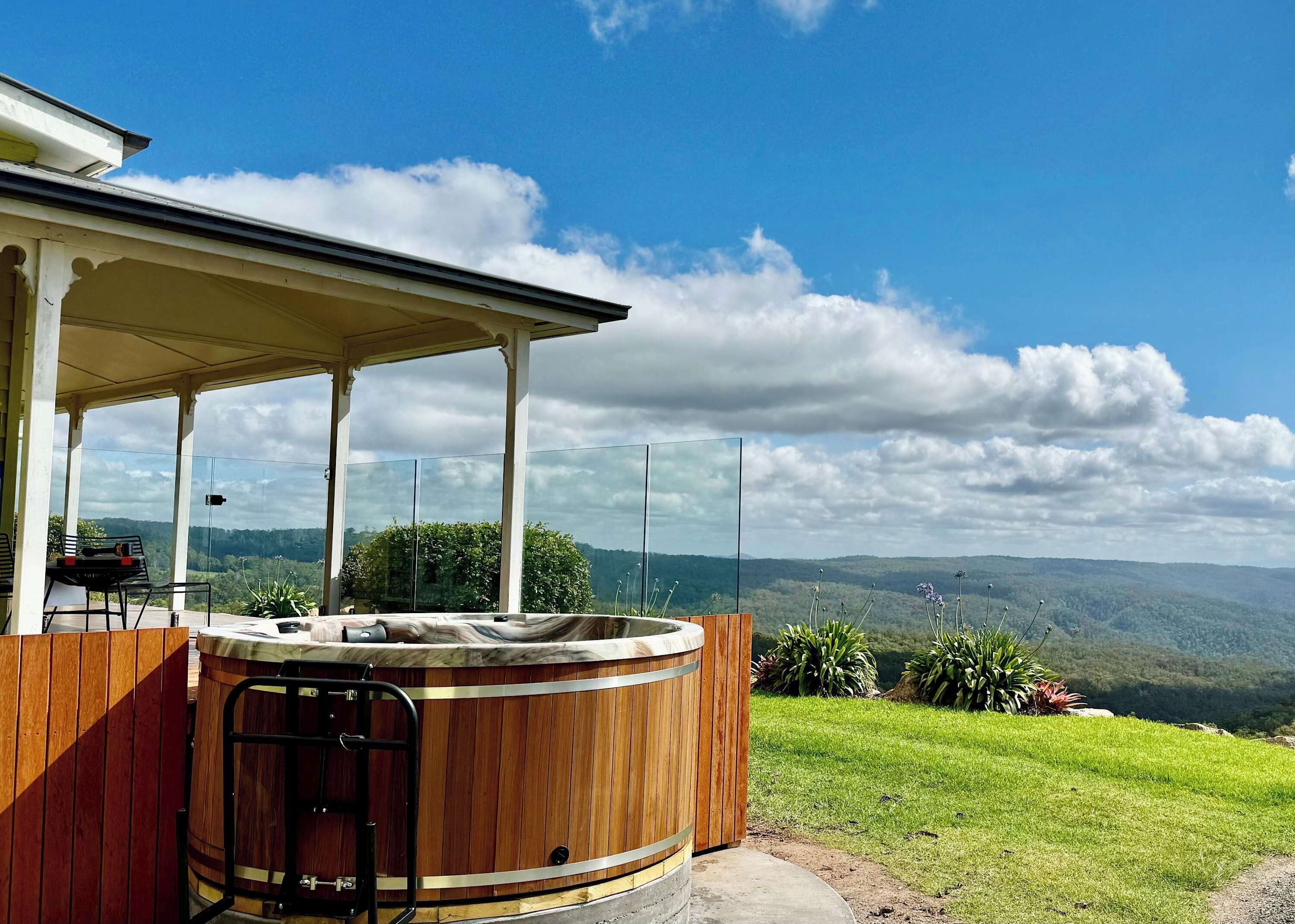 A hot tub with a wooden exterior, a covered patio area, and a scenic view of hills and clouds.