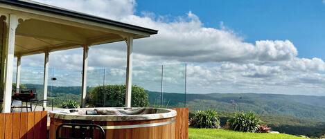 A hot tub with a wooden exterior, a covered patio area, and a scenic view of hills and clouds.