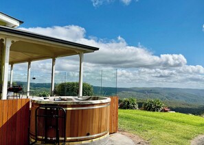 A hot tub with a wooden exterior, a covered patio area, and a scenic view of hills and clouds.