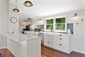 A modern kitchen with white cabinetry, a central island, a stove, and a window with a view of greenery. - The Quince Cottage - 3 Bedrooms (Ravensbourne)