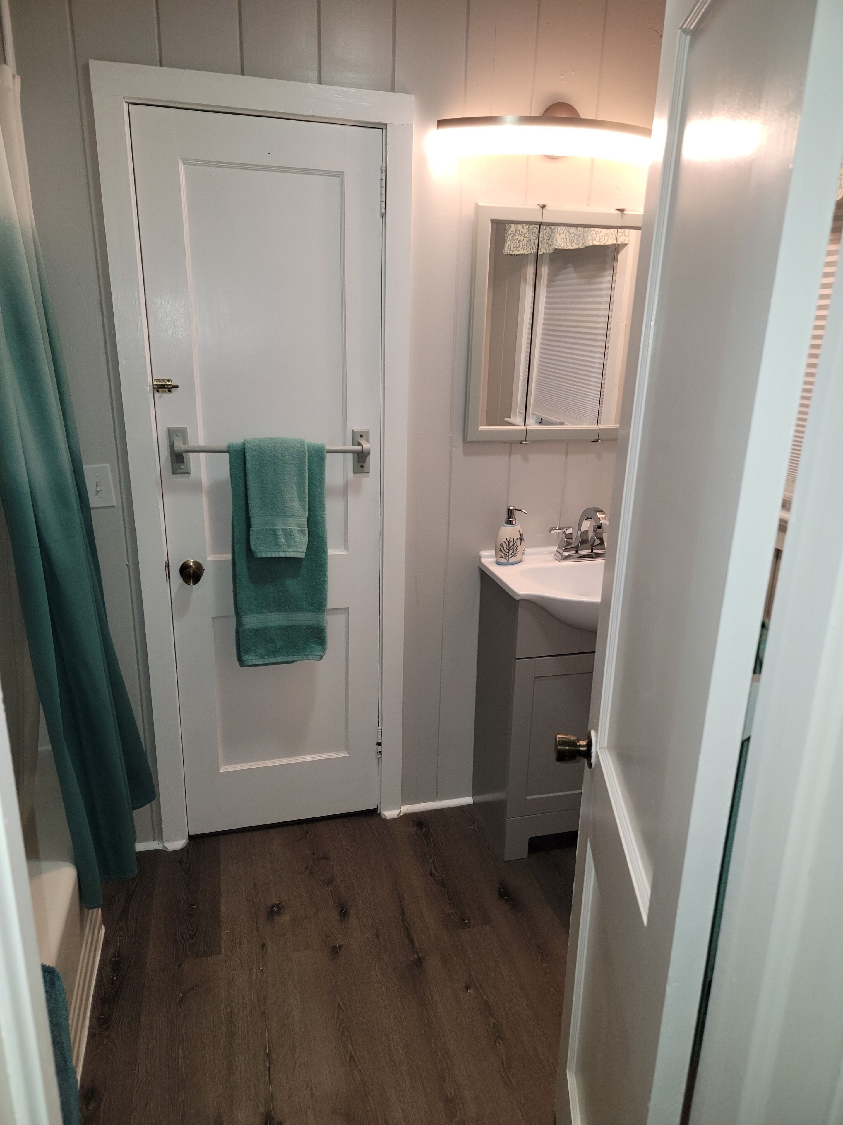 Une salle de bain avec une porte blanche, une serviette turquoise accrochée à la poignée de la porte, un lavabo blanc avec un miroir au-dessus, et un plancher en bois.