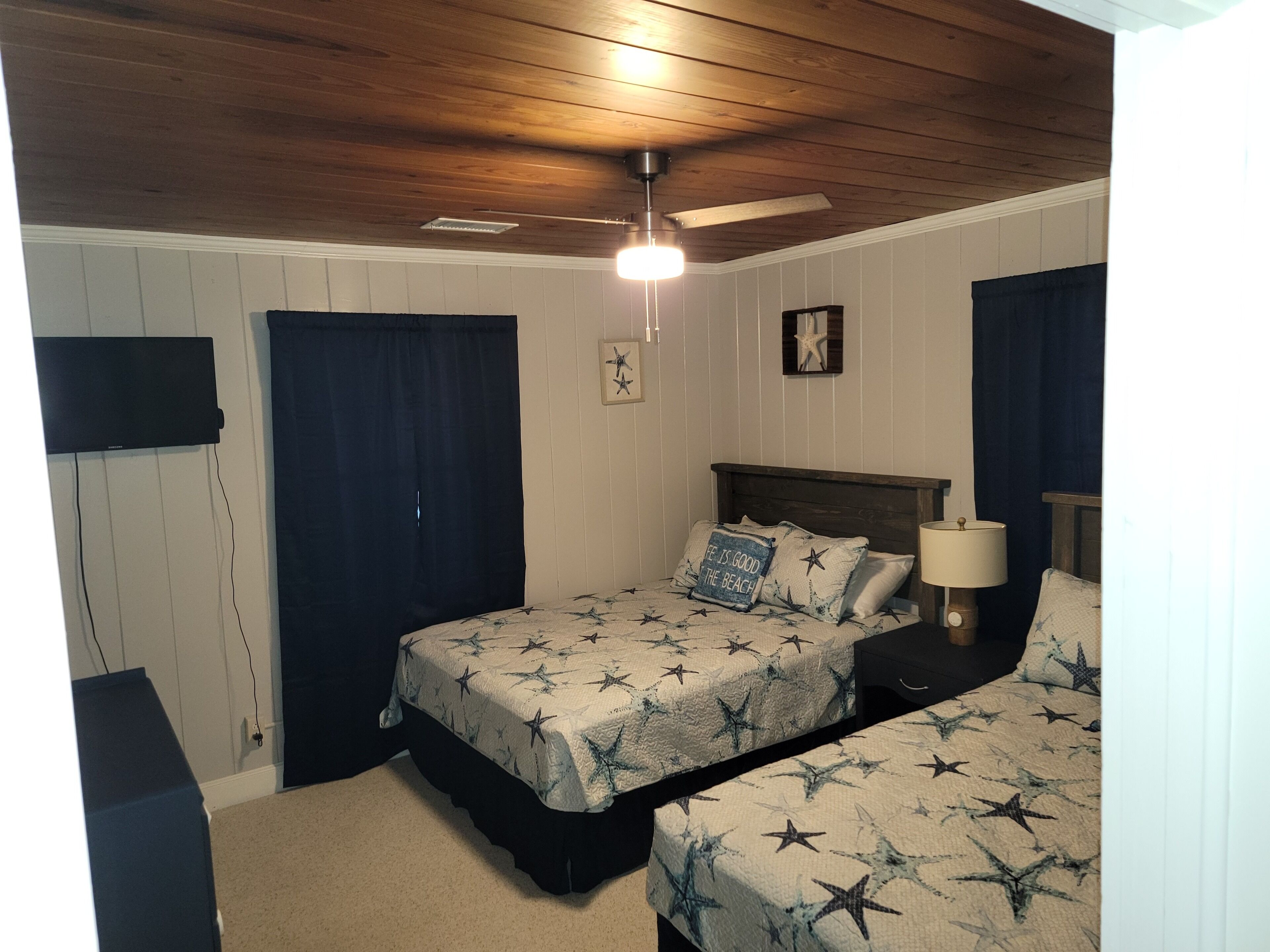 Un dormitorio con dos camas, ventilador de techo, televisor montado en la pared y decoración con forma de estrella de mar.