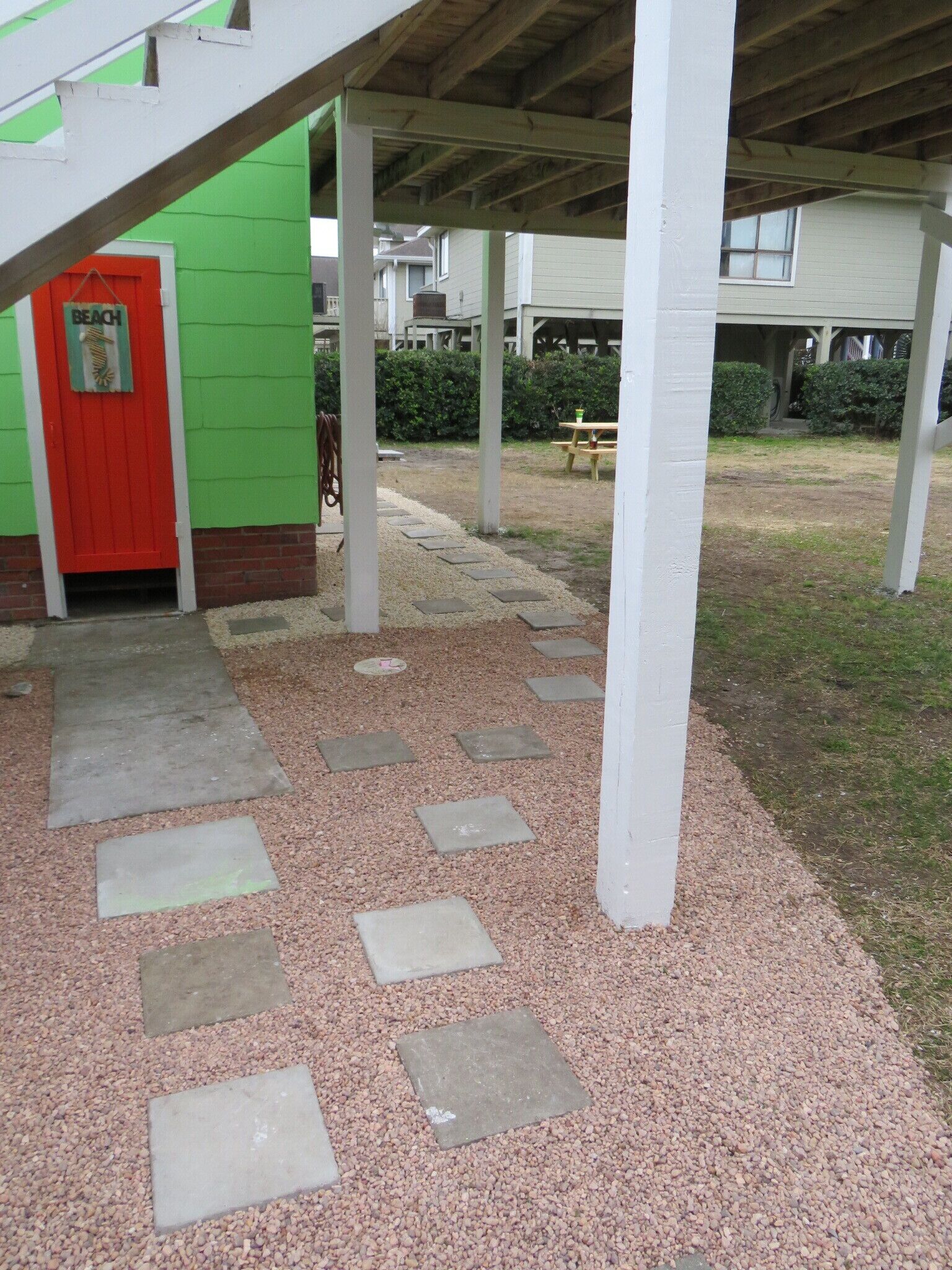 Uma área externa coberta com um caminho de cascalho que leva a uma porta vermelha com a inscrição 'BEACH'.
