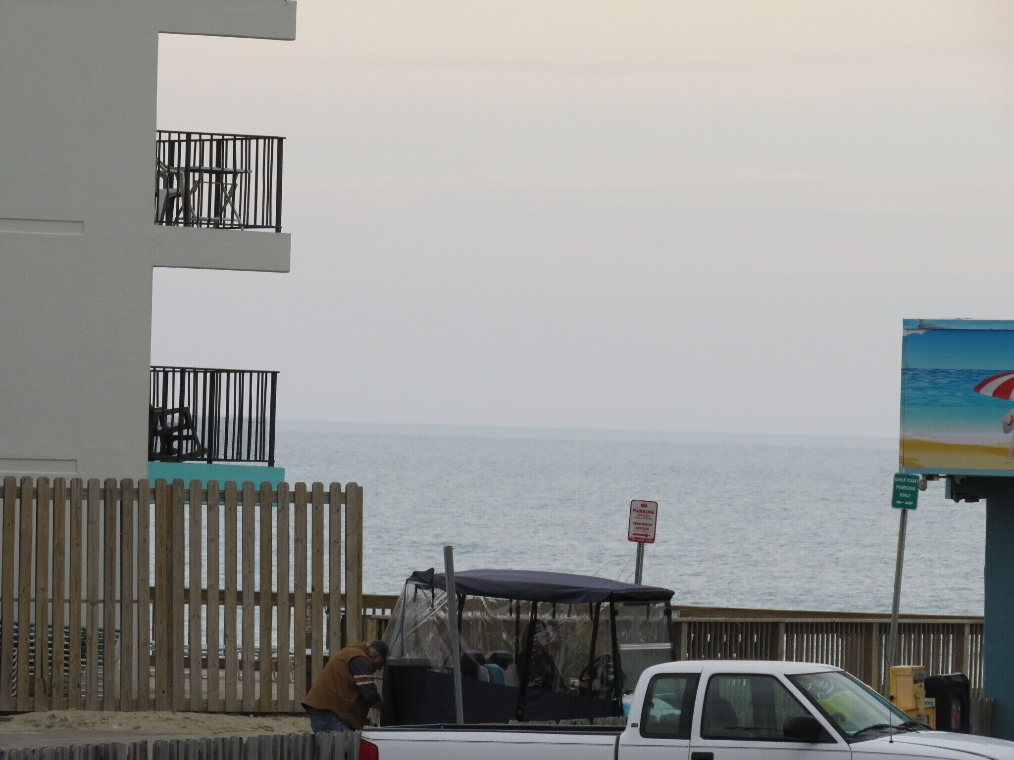A beachside view with a white pickup truck parked near a wooden fence, a person standing by the truck, and a sign indicating 'GOLF CART PARKING ONLY'.