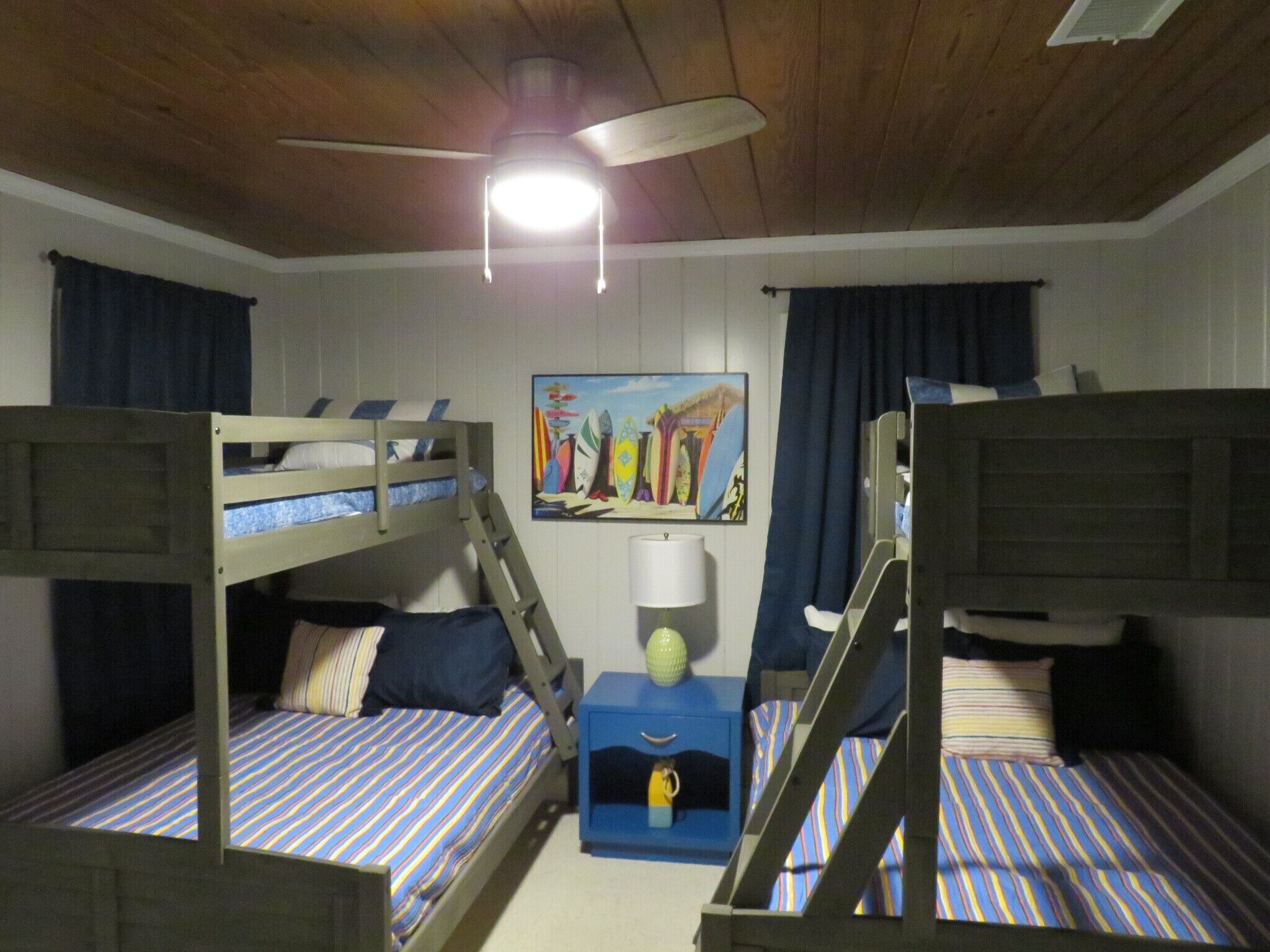 A room with bunk beds, a ceiling fan, a painting, and a small table with a lamp.