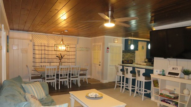 A cozy living room with a sofa, a dining table with chairs, a kitchen area with a bar, and a ceiling fan. - Location Location Location! Classic Beach Cottage less than 1-min walk to beach! (Murrells Inlet)
