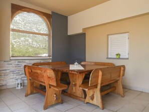 Une table à manger en bois, accompagnée de bancs, se trouve dans une pièce aux murs en pierre, dotée d’une grande fenêtre en arc de cercle et agrémentée d’une petite plante en pot.