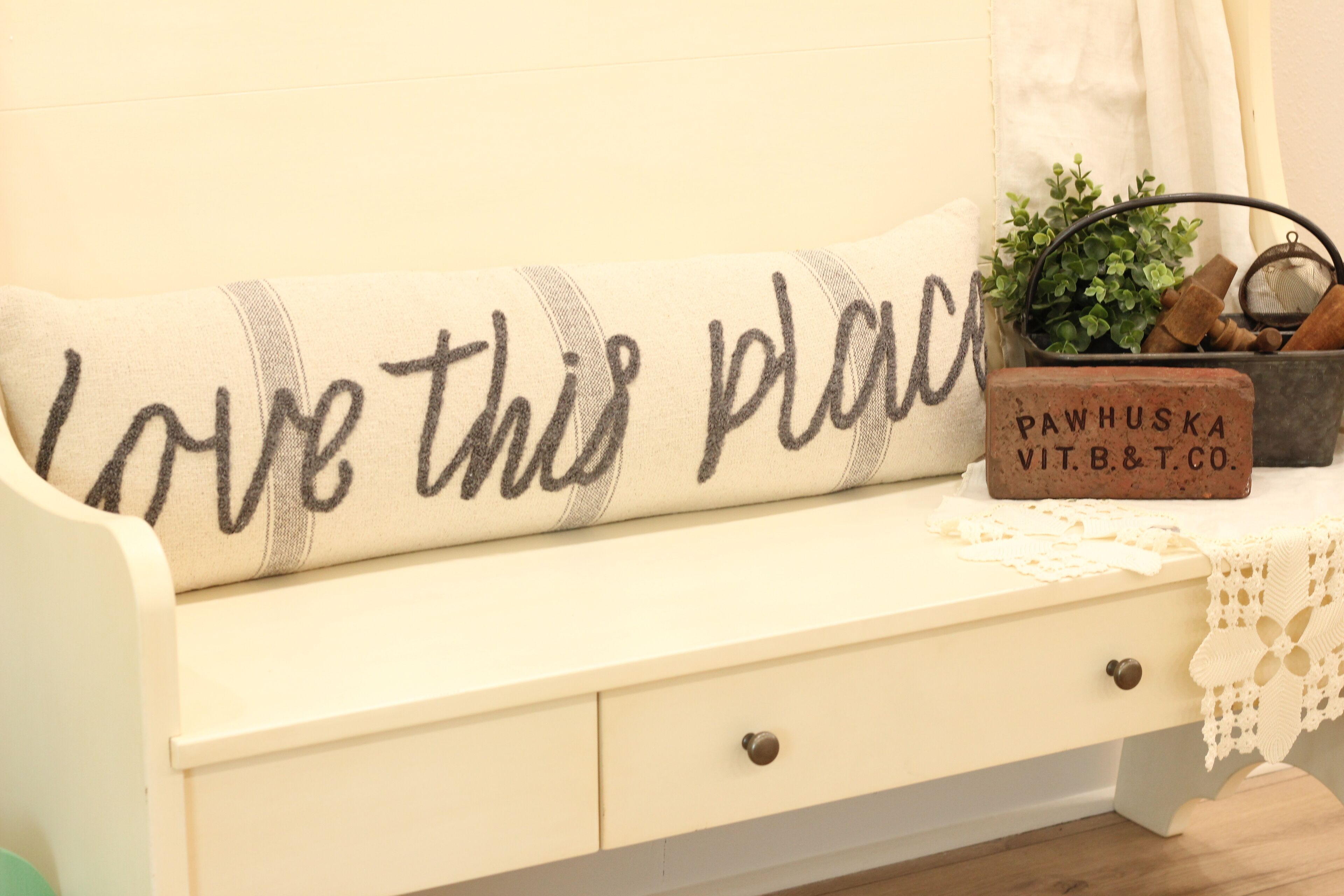 Un sofá blanco con un cojín que dice “Love this place”. Una planta pequeña y un bloque de madera con “PAWHUSKA VIT.B.&T.CO.” están sobre la mesa junto al sofá.