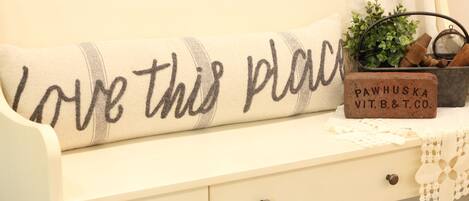 Un sofá blanco con un cojín que dice “Love this place”. Una planta pequeña y un bloque de madera con “PAWHUSKA VIT.B.&T.CO.” están sobre la mesa junto al sofá.