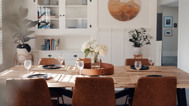 Un comedor con una mesa de madera, sillas tapizadas en tono café y un jarrón con flores blancas.