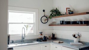 Una cocina con ventana, armarios blancos, un fregadero, una estufa y estantes con utensilios de cocina.