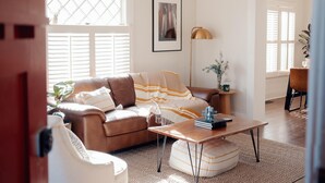 Un salón con un sofá de cuero marrón, una mesa de centro de madera y un sillón blanco. Hay un cuadro en la pared y una lámpara de pie.