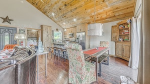 A spacious living area with a wooden ceiling, a dining table, a floral armchair, and a sofa.