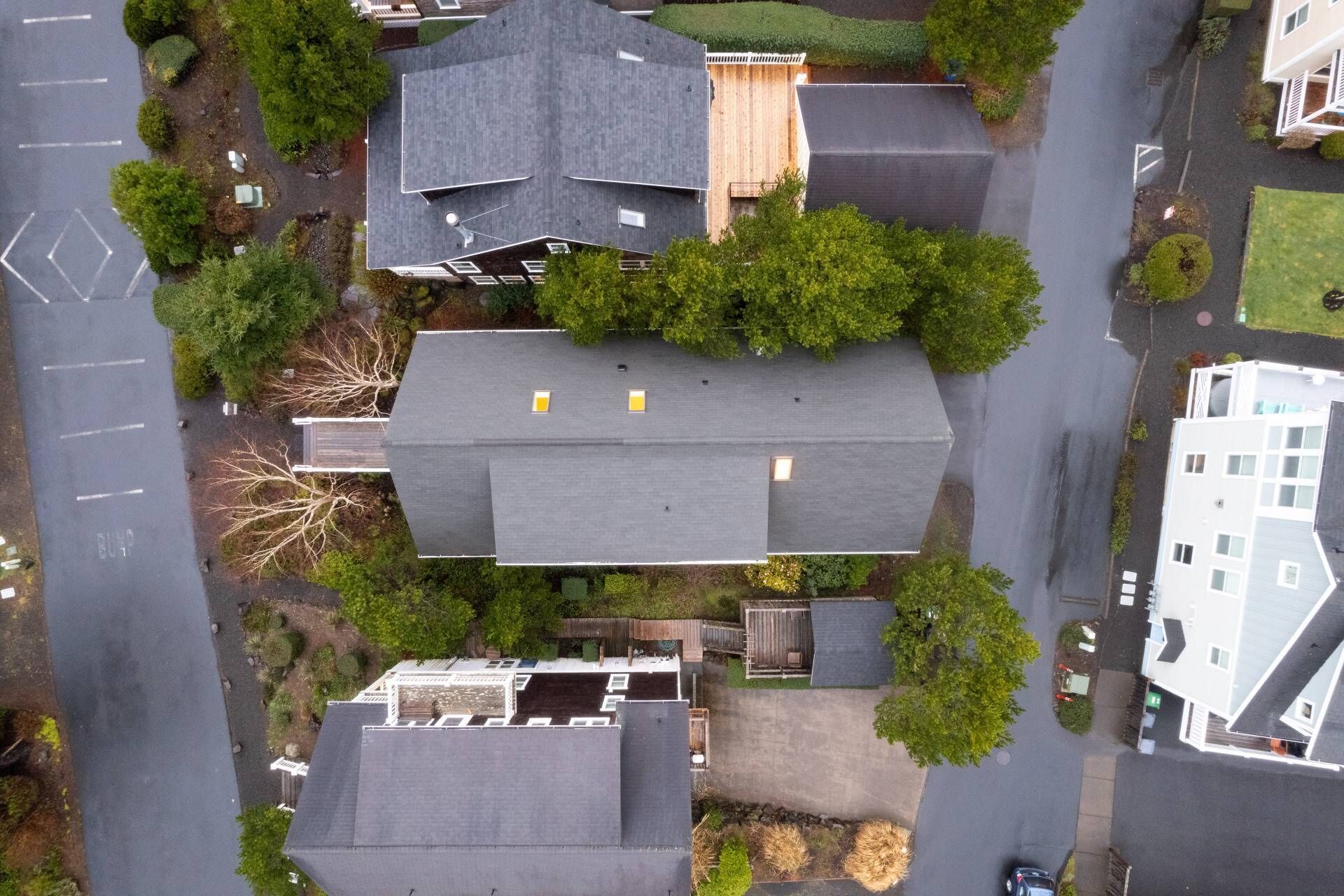 Ett flygbildsbild av ett bostadsområde med flera hus, vart och ett med unika takformer och -storlekar, omgivet av grönska och en huvudväg.