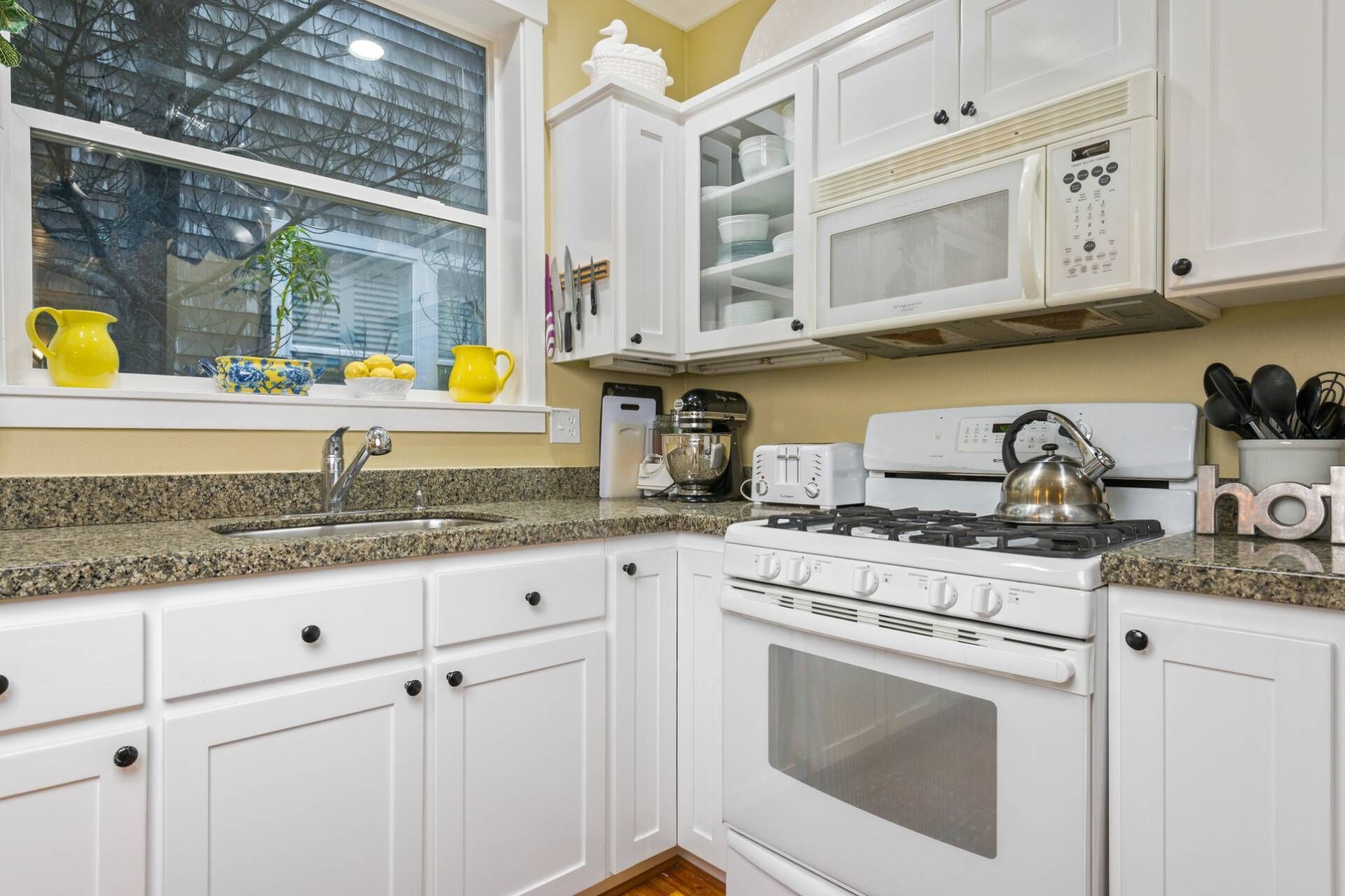 Una cocina con armarios blancos, encimera de granito, estufa, microondas y ventana con jarras amarillas.