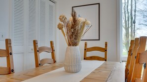 Una mesa de comedor con un jarrón de flores secas, sillas de madera y un cuadro en la pared.
