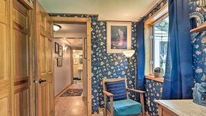 Een kamer met een houten deur, een schommelstoel, bloemenbehang, een lijst met een afbeelding en een raam met gordijnen.
