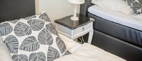 Un lit bien fait, avec un oreiller blanc et noir à motifs de feuilles, une table de chevet avec une lampe et une tête de lit grise.