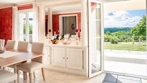 A dining area with a wooden table and chairs, a white cabinet, and a view of a green landscape through a glass door. - Nice villa with private pool and pool house (Montaigu de Quercy)