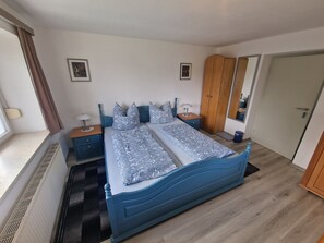 Un dormitorio con cama doble, mesita de noche, armario de madera, espejo y ventana con cortinas.