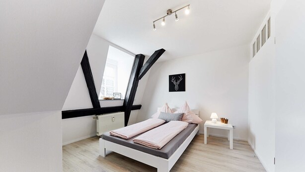 Een moderne slaapkamer met een schuine kap, een bed met een grijze matras en roze beddengoed, een nachtkastje met een lamp, en een afbeelding van een hertenhoofd in een lijst aan de muur.