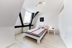 Une chambre moderne avec un plafond en pente, un lit doté d’un matelas gris et de draps roses, une table de chevet avec une lampe, et une œuvre d’art représentant une tête de cerf encadrée au mur.