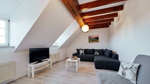 Een moderne loft-woonkamer met een schuine kap, houten balken, een flatscreen televisie, een grijze bank en een kleine salontafel.