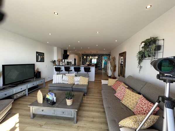 A modern living room with a large flat-screen TV, a grey sofa, a coffee table with decorative items, and a kitchen in the background.