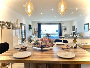A modern dining area with a wooden table set for a meal, a kitchen in the background, and a view of the sea through large windows. - Pleasant, comfortable apartment with sea view (Granville)