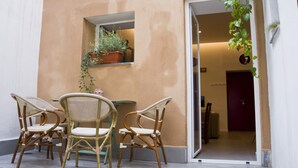 Un petit espace extérieur aménagé avec des chaises en osier et une table, une plante en pot posée sur une étagère, et une porte donnant sur un espace intérieur.