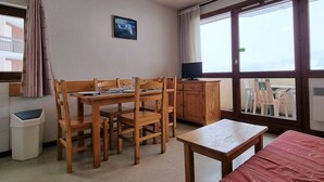 Una habitación acogedora con una mesa y sillas de madera, un pequeño armario, una cama individual, una ventana con cortinas y una lámpara colgante.