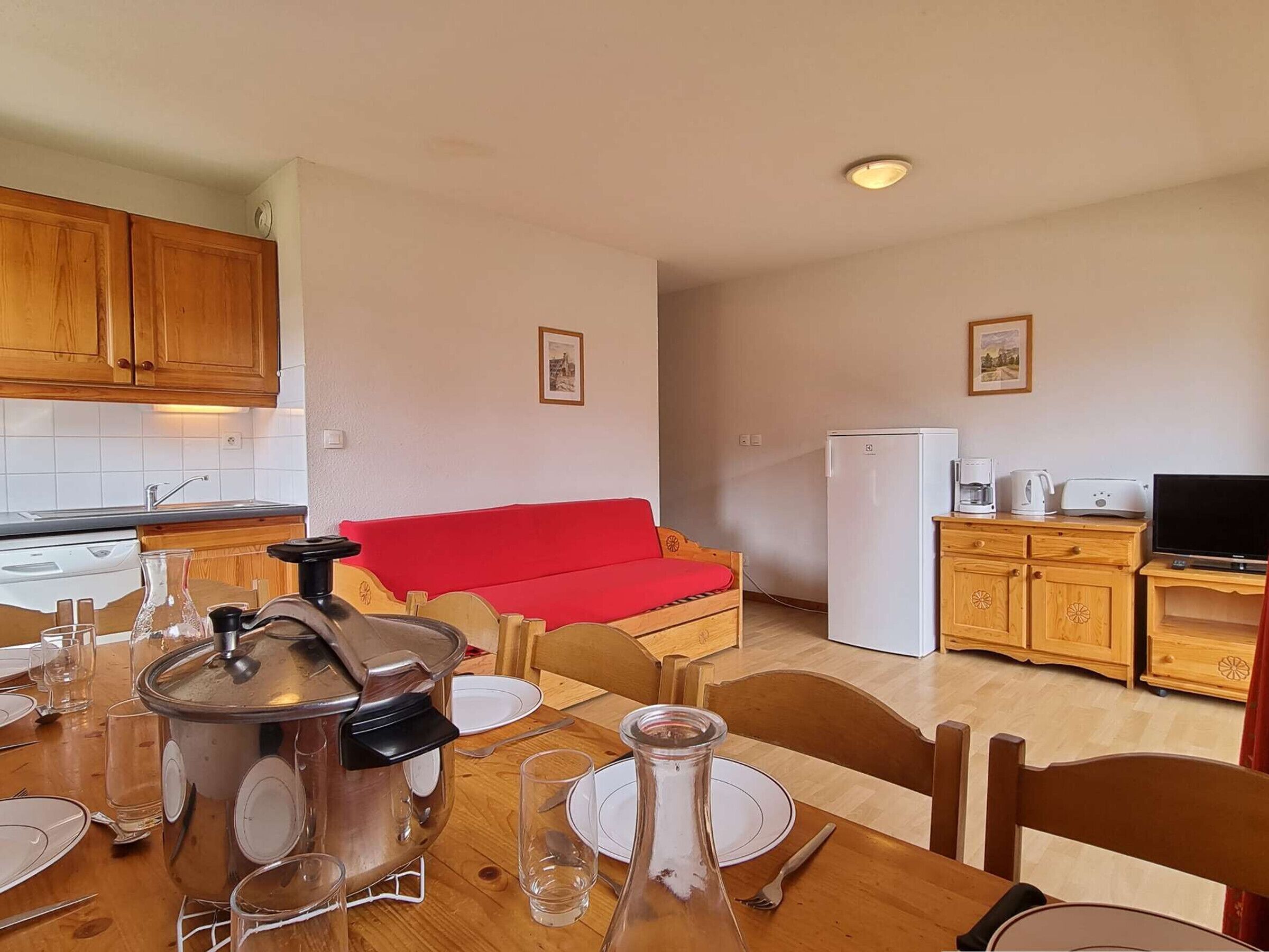 A compact kitchen with wooden cabinets, a red sofa, a white refrigerator, and a dining table set for four.