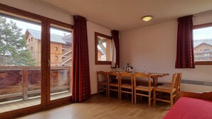 Een kamer met een houten eettafel en stoelen, grote ramen met gordijnen, en een rood tapijt.