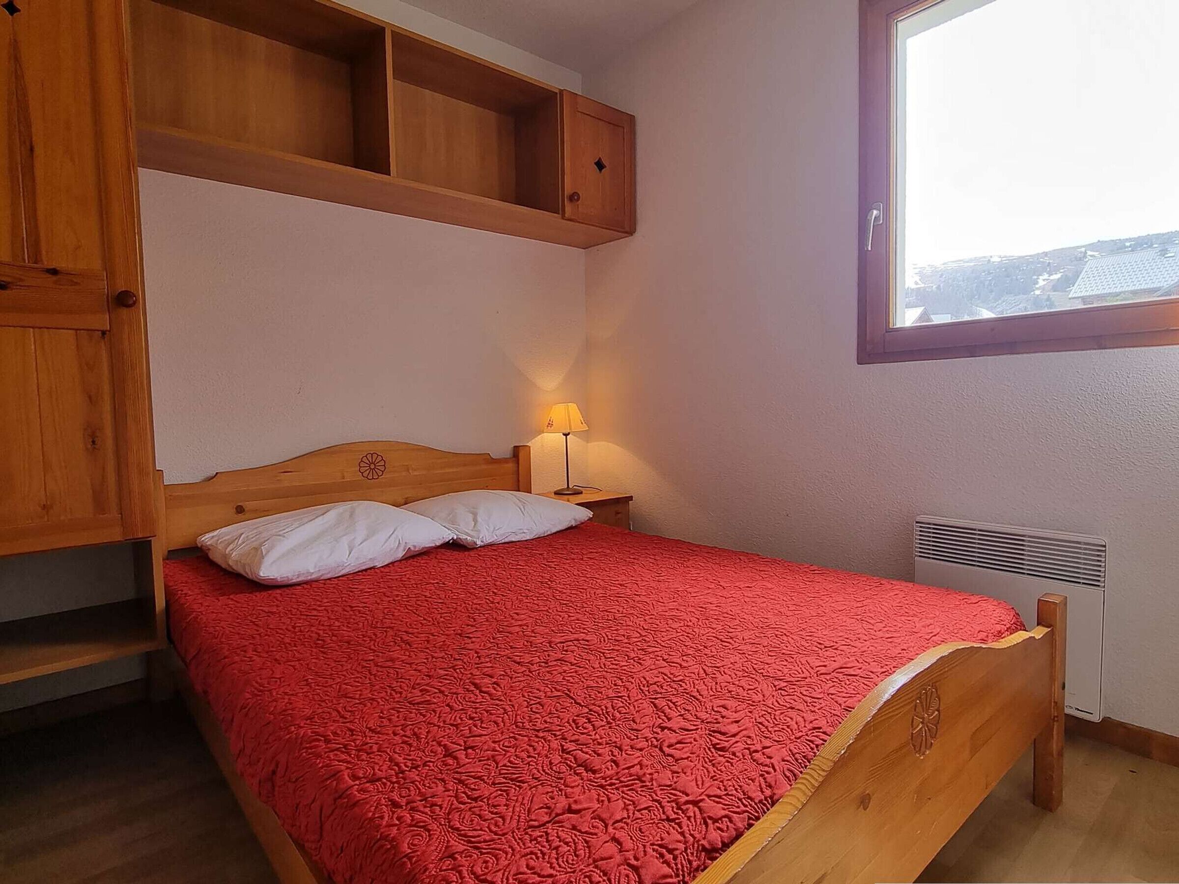 Een slaapkamer met een houten bed, een rode sprei, een nachtkastje met een lamp en een raam met uitzicht op gebouwen.