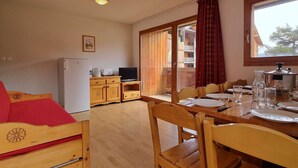 Een gezellige kamer met een houten bed, een klein kitchenette, een eettafel gedekt voor vier personen en een groot raam met uitzicht op een gebouw en bomen.